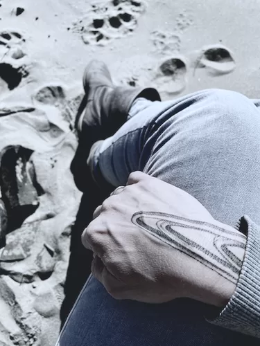 A hand with a graphical tattoo on it, resting on a lap of a person whose legs are in the sand.